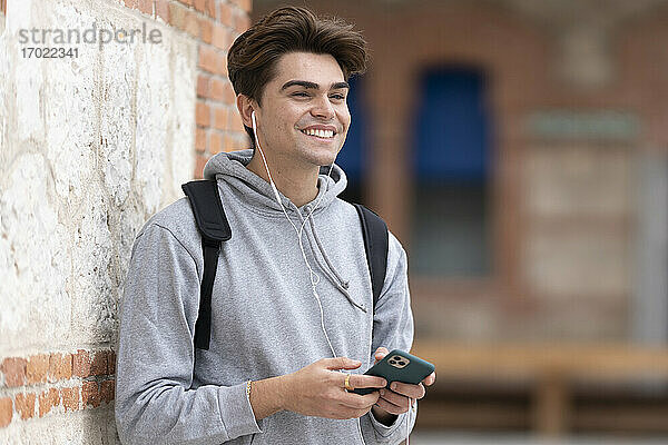 Fröhlicher  gut aussehender junger Mann hört Musik und hält sein Smartphone an die Wand