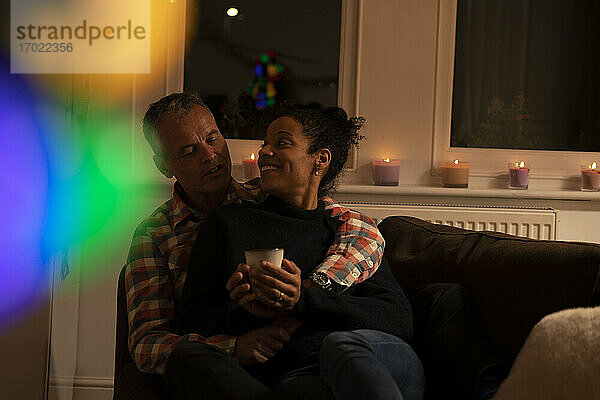 Lächelnde Frau hält Tasse  während sie sich mit ihrem Mann auf dem Sofa zu Hause entspannt