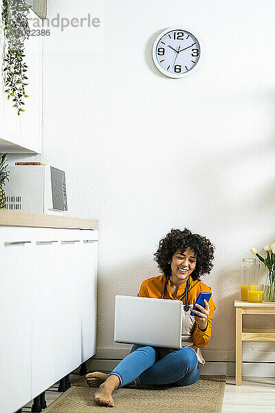 Junge Frau mit Laptop  die lächelt  während sie ein Mobiltelefon benutzt und zu Hause sitzt