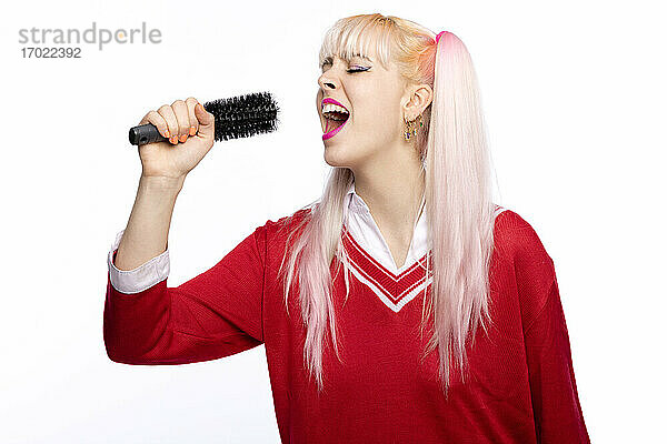 Junge Frau singt durch eine Haarbürste als Mikrofon vor weißem Hintergrund