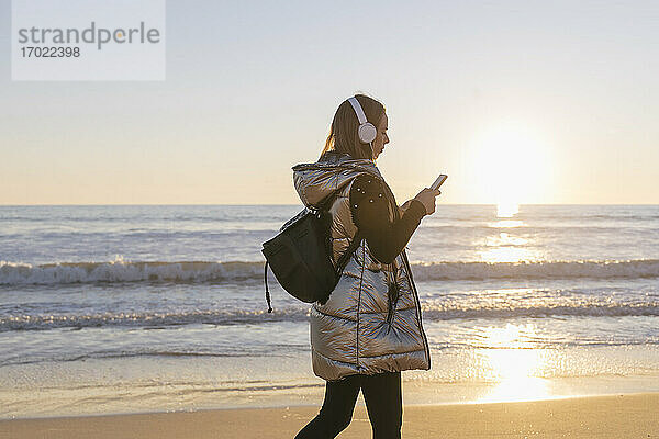 Frau  die Musik hört und ihr Smartphone benutzt  während sie am Strand bei Sonnenuntergang spazieren geht