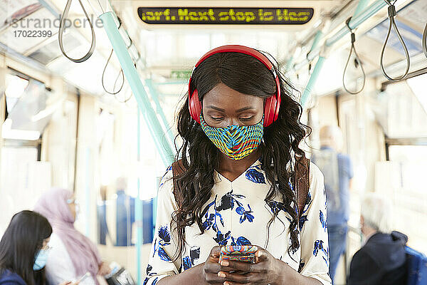 Geschäftsfrau mit Gesichtsmaske und Kopfhörer  die im Zug stehend ein Mobiltelefon benutzt