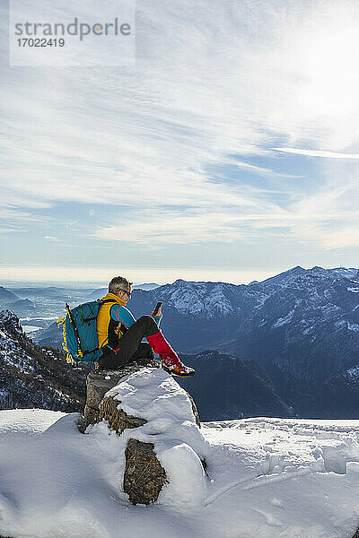 Älterer Mann mit Rucksack  der ein Mobiltelefon benutzt  während er auf einem verschneiten Berg sitzt  Orobic-Alpen  Lecco  Italien