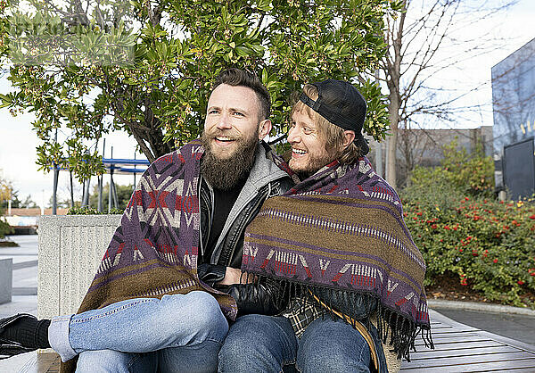 Fröhliche schwule Freunde  eingewickelt in einen Schal  sitzen auf einer Bank