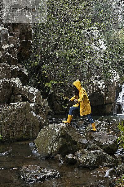 Frau in gelbem Regenmantel klettert auf Felsen im Bach im Wald