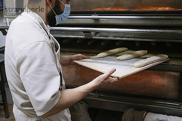Männlicher Koch beim Einschieben von Brotteig in den Ofen in einer Bäckerei während COVID-19