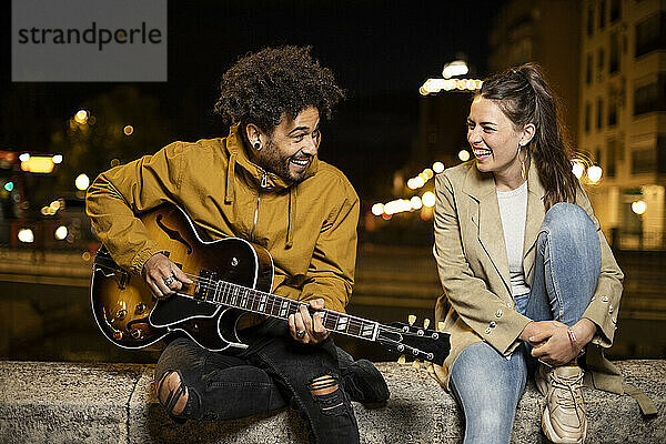 Glückliches Paar mit Gitarre auf Stützmauer in der Stadt sitzend