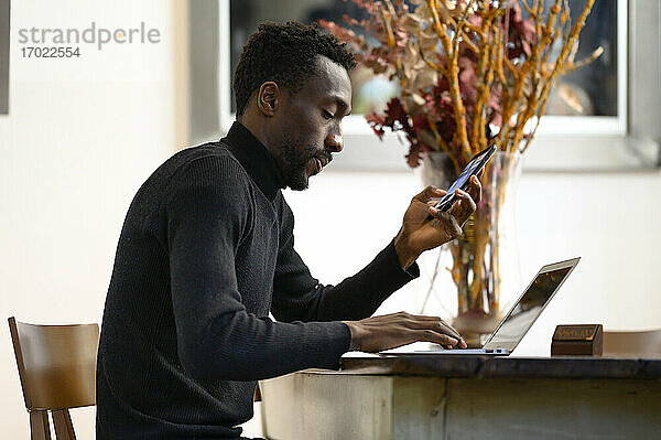 Junger Geschäftsmann mit Mobiltelefon  der in einem Café sitzt und am Laptop arbeitet