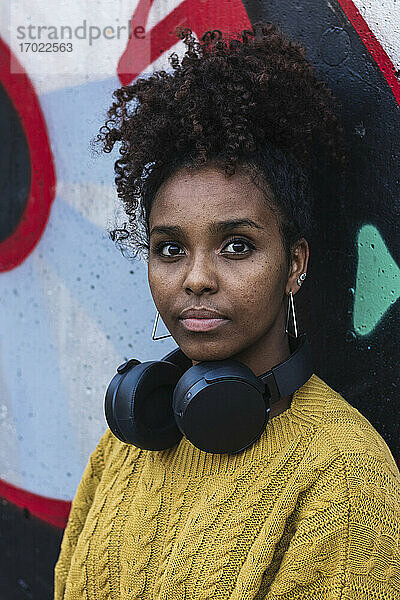 Selbstbewusste junge Frau mit lockigem Haar und Kopfhörern vor einer Graffiti-Wand