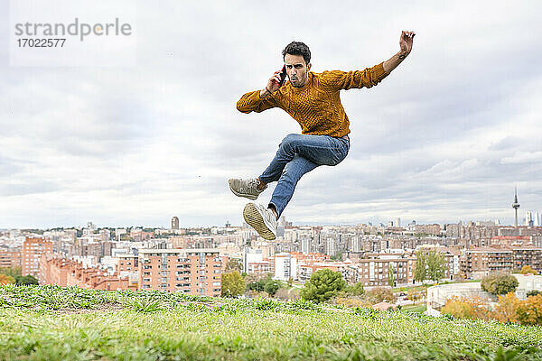 Junger Mann springt während des Telefonierens über einen Hügel vor einer Stadtlandschaft