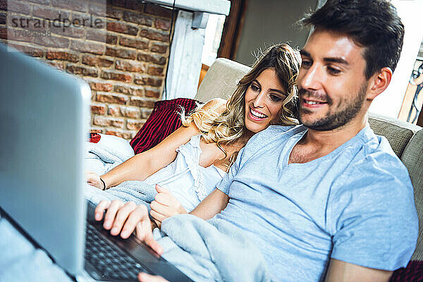 Glückliches junges Paar sitzt zu Hause auf der Couch und schaut auf den Laptop