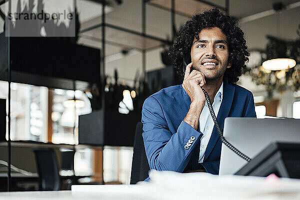 Lächelnder Geschäftsmann  der mit einem Laptop im Büro sitzt und telefoniert