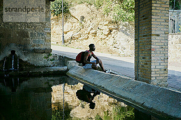 Junger Mann sitzt am Wasser und macht eine Pause beim Spaziergang