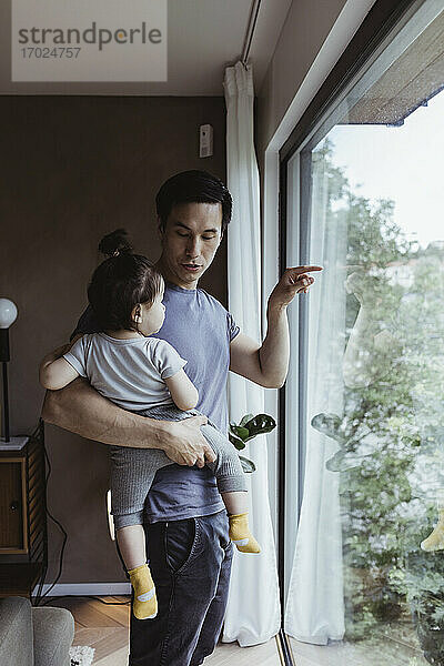Vater  der aus dem Fenster zeigt  während er ein männliches Kleinkind zu Hause trägt