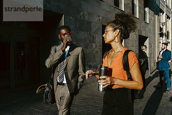 Männlicher Unternehmer trinkt Kaffee  während er mit einer weiblichen Kollegin auf einem Fußweg in der Stadt diskutiert