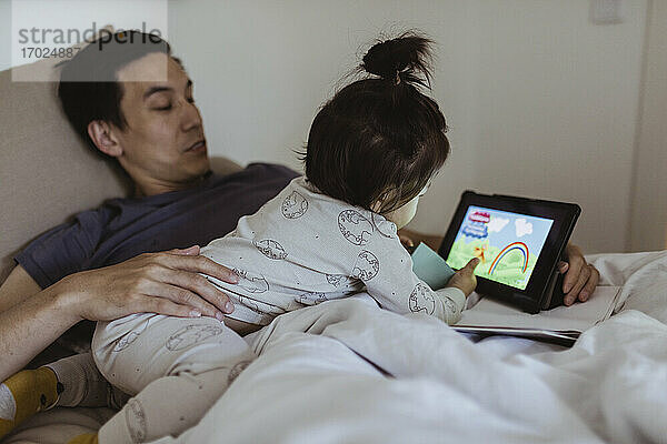 Männliches Kleinkind schaut Cartoon auf digitalem Tablet mit Vater im Schlafzimmer