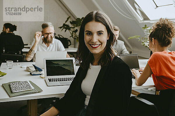 Lächelnde junge Geschäftsfrau sitzt mit Kollegen im Coworking-Büro