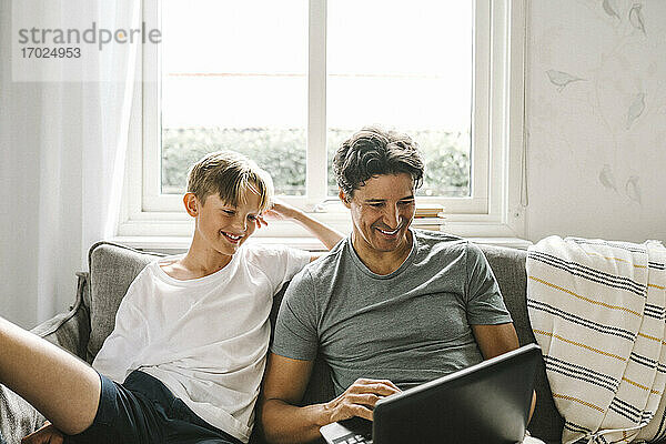 Glücklicher Vater mit Laptop  während er bei seinem Sohn im Wohnzimmer sitzt