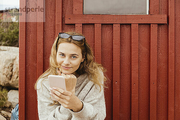 Lächelnde Frau mit Hand am Kinn hält Smartphone gegen Hütte