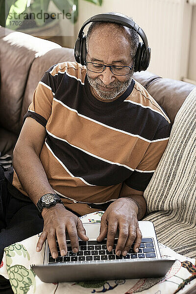 Älterer Mann hört Musik über Kopfhörer  während er einen Laptop im Wohnzimmer benutzt
