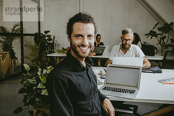 Porträt eines lächelnden männlichen Unternehmers  der im Büro am Laptop sitzt