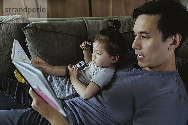 Vater mit männlichem Kleinkind liest Buch  während er auf dem Sofa im Wohnzimmer liegt