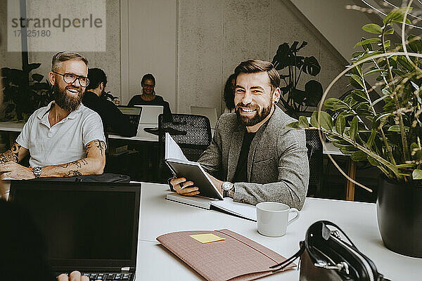 Lächelnde Geschäftsmann hält Tagebuch beim Sitzen mit männlichen Kollegen in Coworking Büro