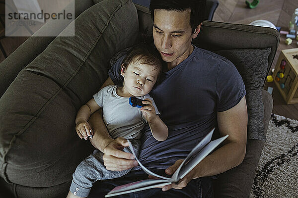 Hohe Winkel Ansicht von Vater mit männlichen Kleinkind Lesen Buch  während auf dem Sofa zu Hause liegen
