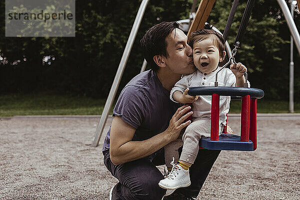 Lächelnder Vater küsst Sohn  während er auf einer Schaukel auf dem Spielplatz sitzt