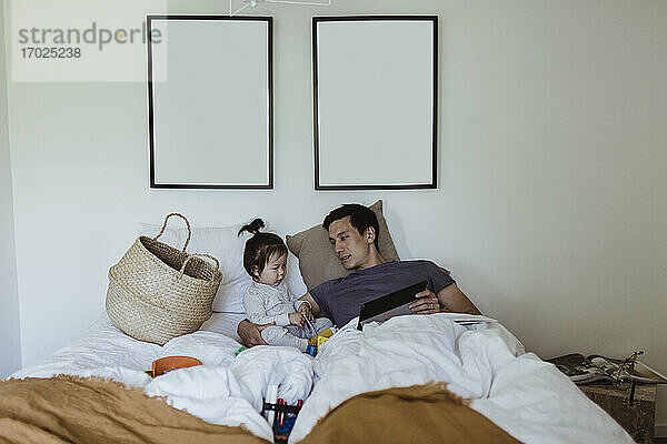 Vater mit männlichem Kleinkind hält digitales Tablet im Schlafzimmer zu Hause