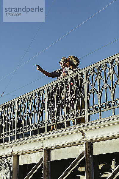 Älterer Mann  der auf eine Frau auf einer Brücke gegen den blauen Himmel zeigt