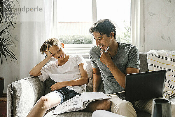 Lächelnder Vater sitzt bei Sohn mit Buch auf Sofa im Wohnzimmer