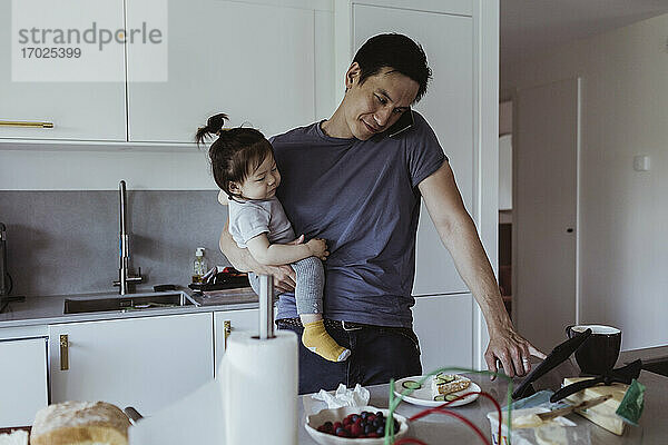 Vater spricht auf Smartphone während der Verwendung von digitalen Tablet  während er Baby Sohn in der Küche trägt