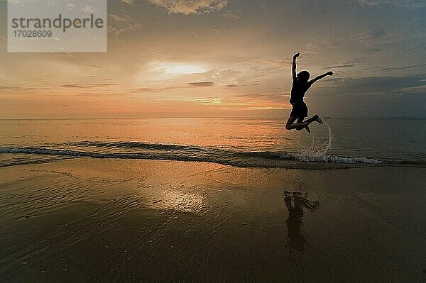 Eine junge Frau springt vor Freude und feiert am Strand bei Sonnenuntergang.