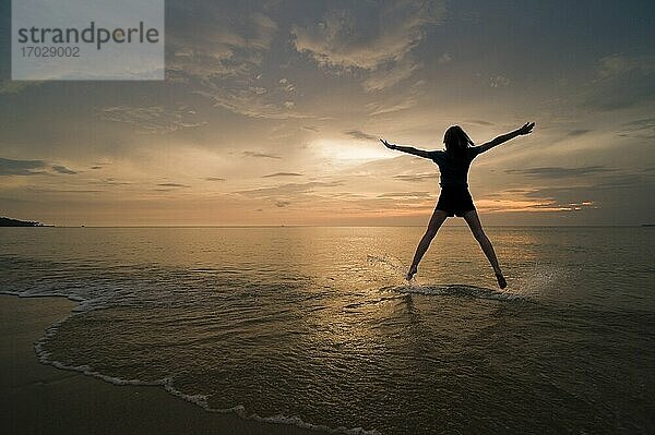 Eine junge Frau macht einen Sternsprung und genießt ihre Freiheit am Strand bei Sonnenuntergang.