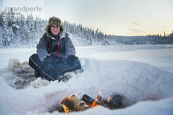 Eine Person sitzt an einem Lagerfeuer und wärmt sich an einem eiskalten Tag im Winter am Polarkreis in Lappland  Finnland