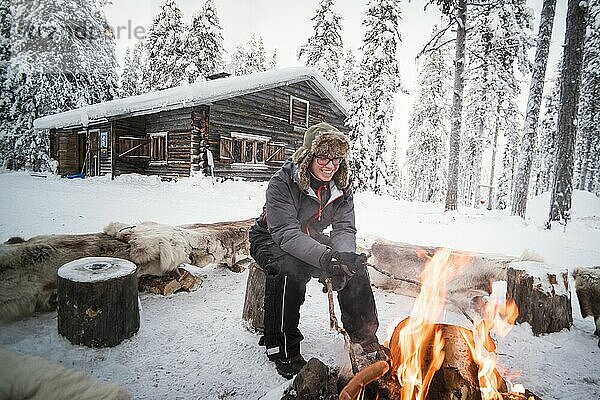 Eine Person sitzt an einem Lagerfeuer und wärmt sich an einem eiskalten Tag im Winter am Polarkreis in Lappland  Finnland
