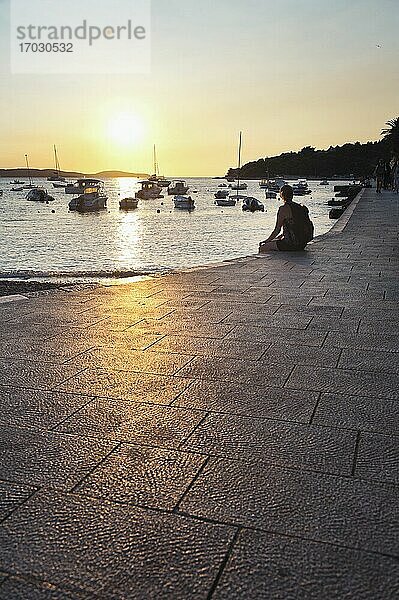 Tourist sitzt und beobachtet den Sonnenuntergang im Hafen der Stadt Hvar  Insel Hvar  Dalmatien  Kroatien. Dies ist ein Foto eines Touristen  der den Sonnenuntergang im Hafen der Stadt Hvar  Insel Hvar  Dalmatien  Kroatien  beobachtet. Es gibt viele Orte  an denen man einen Sonnenuntergang in Hvar beobachten kann  und nur wenige sind besser als der alte Hafen.