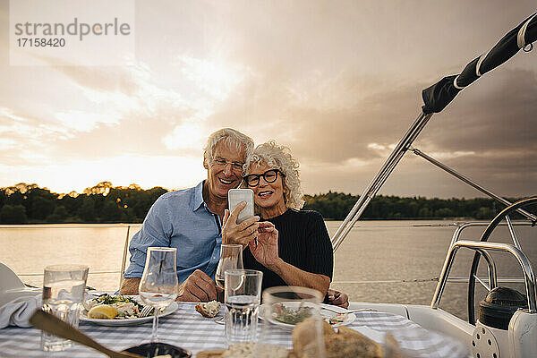 Lächelndes heterosexuelles Paar nimmt Selfie durch Smartphone in Segelboot während Sonnenuntergang