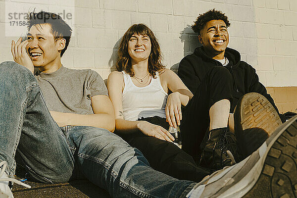 Lächelnde männliche und weibliche Freunde sitzen auf Gehweg gegen Wand während sonnigen Tag
