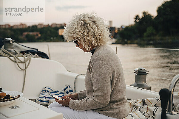 Senior Frau Text-Messaging auf Smartphone  während in Boot während des Sonnenuntergangs sitzen