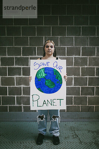 Weiblicher Demonstrant mit Planet-Earth-Plakat steht auf dem Fußweg an der Wand