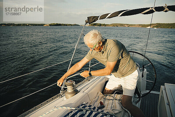 Senior Mann bindet Seil auf Klampe  während kniend in Boot während sonnigen Tag