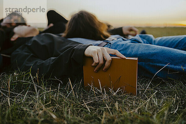 Junger Mann hält Tagebuch liegend von Freunden auf Gras während Sonnenuntergang