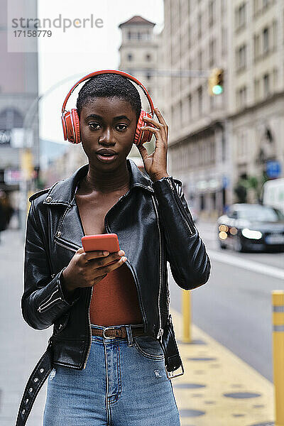 Young woman with mobile phone listening music through headphones while standing in city