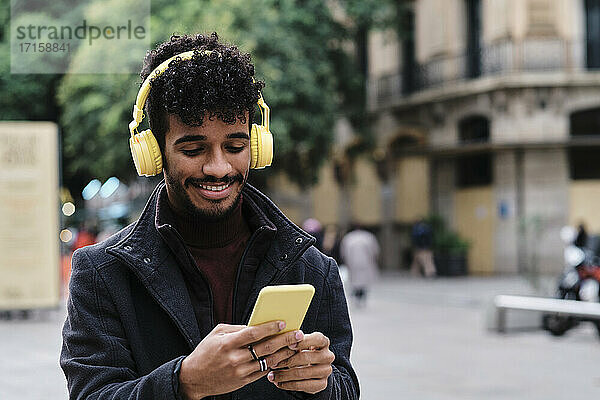 Smiling man wearing headphones using mobile phone while standing in city