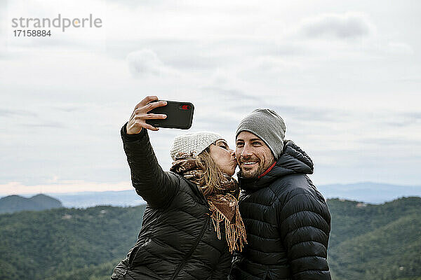 Frau küsst lächelnd Freund während der Einnahme selfie gegen den Himmel im Winter