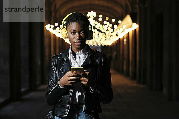 Young woman with headphones and mobile phone staring while standing at corridor