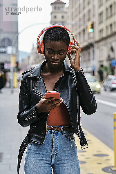Woman listening music through headphones while using smart phone standing in city