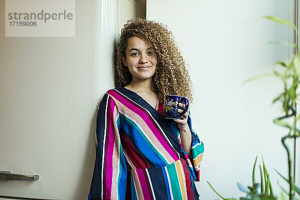 Lächelnde junge Frau  die eine Kaffeetasse hält  während sie an einer weißen Wand im Wohnzimmer steht